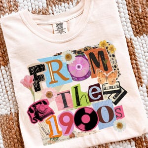 May include: A light peach-colored t-shirt with a collage design that reads "From The 1990s." The design includes a pink rotary phone, flowers, a CD, a cassette tape, a vinyl record, and a butterfly. The shirt is made by Comfort Colors.