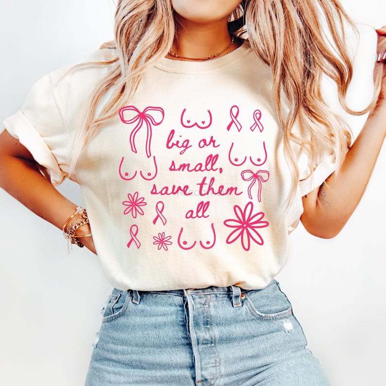 May include: Cream-colored t-shirt with pink text and graphics. The text reads "big or small, save them all" with illustrations of bows, ribbons, flowers, and breasts. The shirt is a casual style.