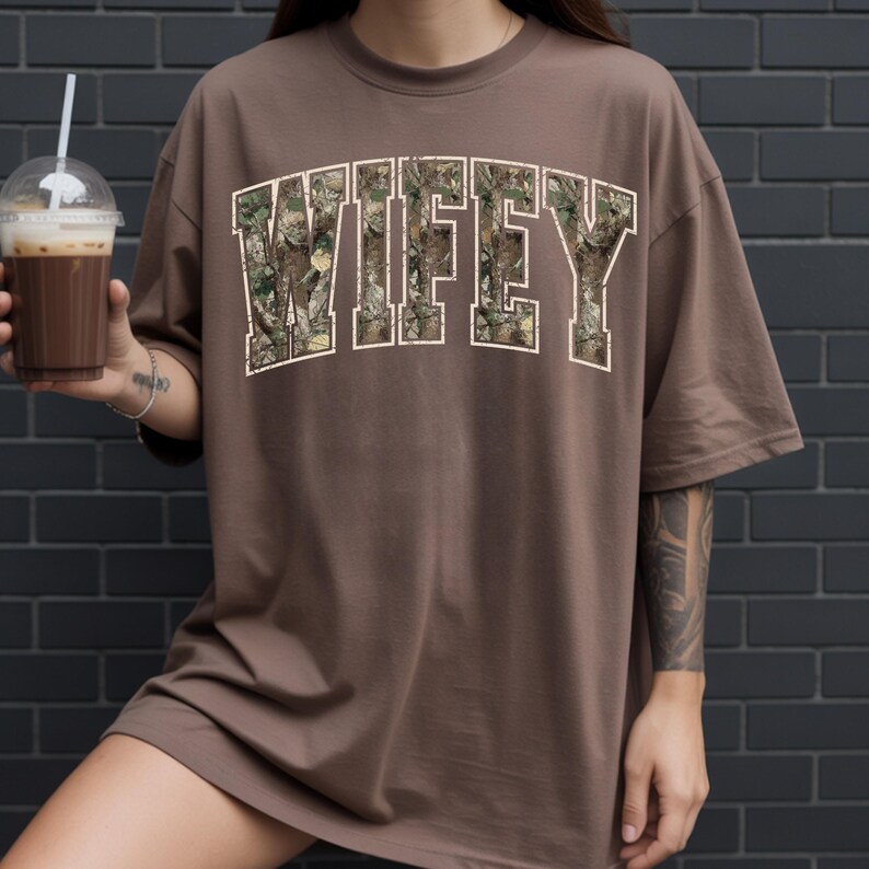May include: A brown t-shirt with the word "WIFEY" in a camouflage pattern. The text is outlined in white. The shirt has short sleeves and a relaxed fit. The image shows a person holding a drink.