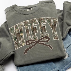 May include: An olive green sweatshirt with the word "WIFEY" in white block letters filled with a camouflage pattern. A brown bow graphic is below the word. The sweatshirt is folded on top of a pair of blue denim shorts.