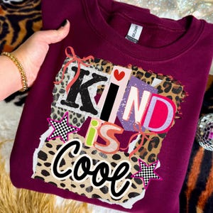 May include: A maroon sweatshirt with the words "KIND is Cool" in a colorful, layered design. The design includes leopard print, stars, and a heart. The sweatshirt is a crew neck style.