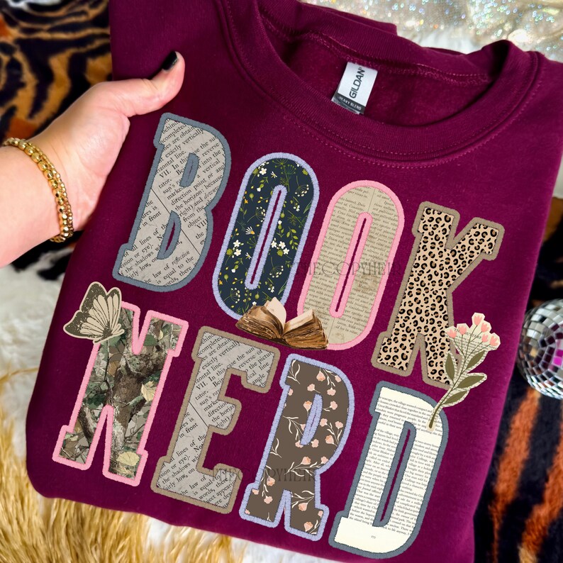 May include: Burgundy sweatshirt with the words "BOOK NERD" in a patchwork design. The letters feature floral, leopard print, and book page patterns. A butterfly and a small open book are also included in the design.
