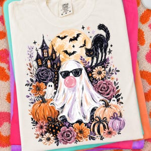 May include: A cream-coloured t-shirt featuring a graphic print of a ghost wearing black sunglasses and blowing a pink bubble. The design includes a haunted house, a black cat, bats, a full moon, pumpkins, and various flowers in shades of purple and orange.