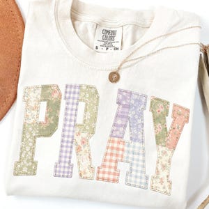 May include: Cream-colored t-shirt with the word "PRAY" appliqued in patchwork floral and gingham fabrics. A gold necklace with a pendant rests on the shirt. The shirt has a Comfort Colors tag.