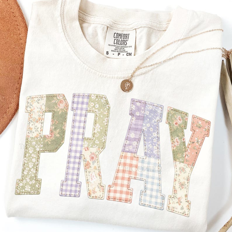 May include: Cream-colored t-shirt with the word "PRAY" appliqued in patchwork floral and gingham fabrics. A gold necklace with a pendant rests on the shirt. The shirt has a Comfort Colors tag.