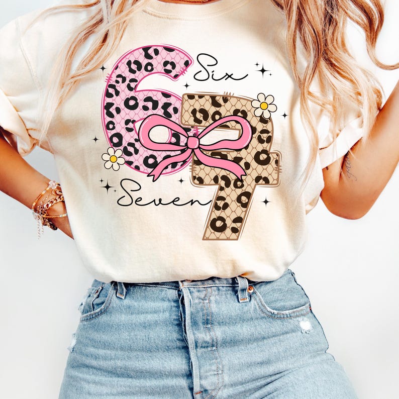 Pode incluir: Camiseta creme com os n&uacute;meros "6" e "7" em um design l&uacute;dico. O "6" &eacute; rosa com estampa de leopardo, enquanto o "7" &eacute; bege com estampa de leopardo. Um la&ccedil;o rosa conecta os n&uacute;meros, com as palavras "Six" e "Seven" em escrita.