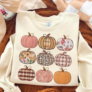 May include: Cream-colored sweatshirt featuring nine illustrated pumpkins with various patterns, including plaid, floral, and leopard print. The pumpkins are in shades of pink, orange, and brown, perfect for autumn.