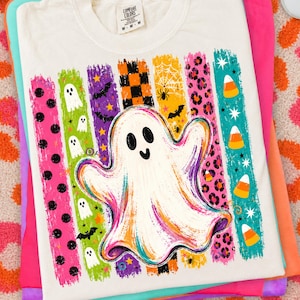 May include: A white t-shirt featuring a large central ghost graphic surrounded by vertical stripes in various colours and patterns, including polka dots, ghosts, bats, checkered squares, spider webs, leopard print, and candy corn icons.