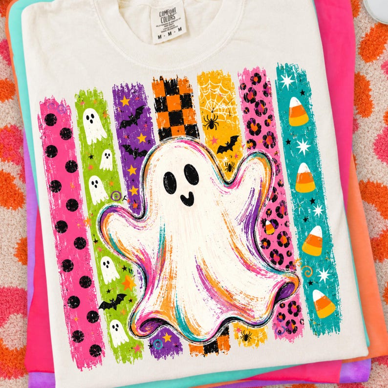 May include: A white t-shirt featuring a large central ghost graphic surrounded by vertical stripes in various colours and patterns, including polka dots, ghosts, bats, checkered squares, spider webs, leopard print, and candy corn icons.