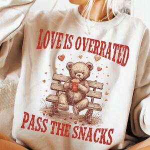 May include: A cream-colored sweatshirt with the text "LOVE IS OVERRATED" and "PASS THE SNACKS" in red. A teddy bear is seated on a park bench, holding a container of popcorn. Small red hearts are scattered around the bear.