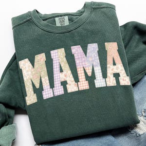 May include: A dark green sweatshirt with the word "MAMA" in large, patchwork letters. Each letter features a different floral or gingham pattern in pastel colors. The sweatshirt has a crew neck and long sleeves.