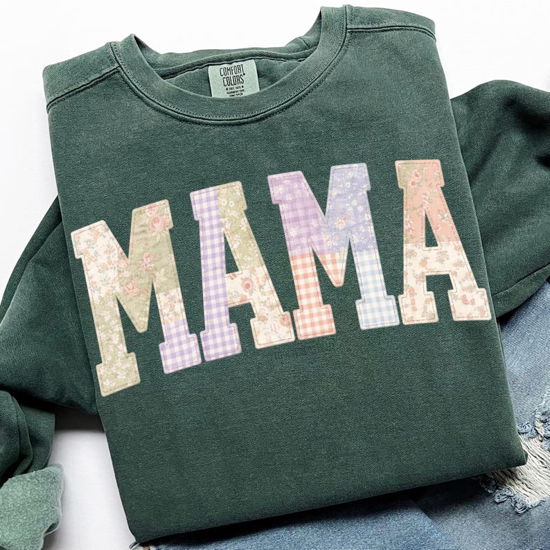 May include: A dark green sweatshirt with the word "MAMA" in large, patchwork letters. Each letter features a different floral or gingham pattern in pastel colors. The sweatshirt has a crew neck and long sleeves.