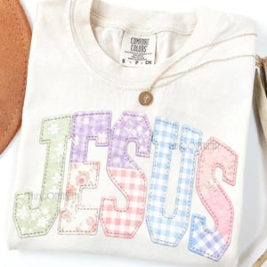 Puede incluir: Camiseta color crema con la palabra "JESUS" aplicada en patrones florales y cuadros vichy pastel. Un collar de oro con un colgante descansa sobre la camiseta. La camiseta tiene una etiqueta Comfort Colors.