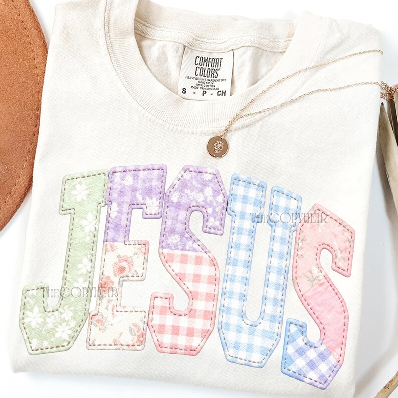 Puede incluir: Camiseta color crema con la palabra "JESUS" aplicada en patrones florales y cuadros vichy pastel. Un collar de oro con un colgante descansa sobre la camiseta. La camiseta tiene una etiqueta Comfort Colors.