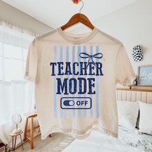 May include: Cream-colored t-shirt with the words "TEACHER MODE" in navy blue, accented by light blue vertical stripes and a bow graphic. The shirt features an "OFF" switch graphic, perfect for educators.
