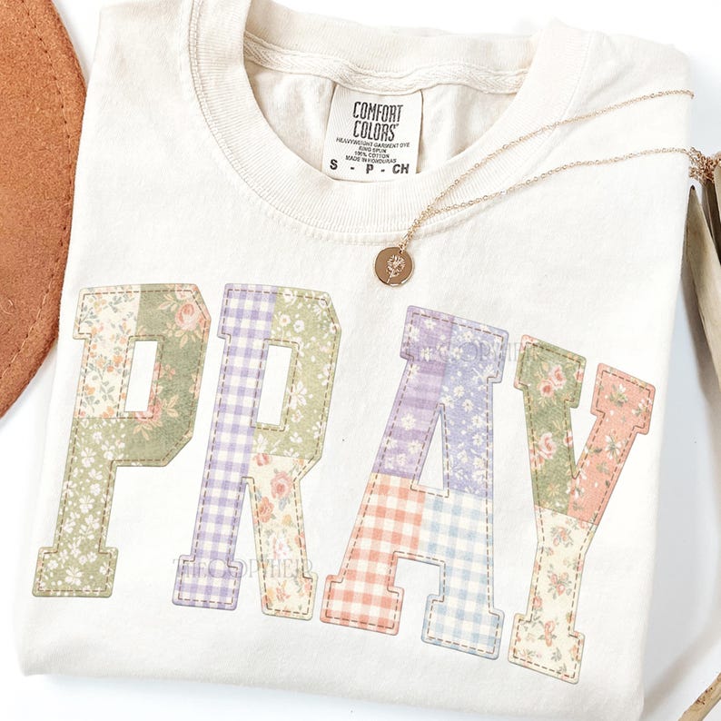 K&ouml;nnte beinhalten: Cremefarbenes T-Shirt mit dem Wort "PRAY" in Patchwork-Buchstaben. Die Buchstaben zeigen Blumen- und Vichy-Muster in Pastellfarben. Eine goldene Halskette mit einem kleinen Anh&auml;nger liegt auf dem Shirt.