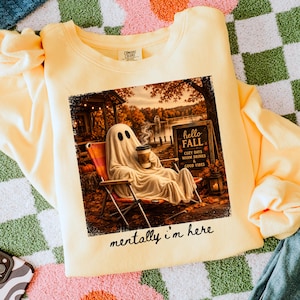 May include: A light yellow sweatshirt with a graphic of a ghost relaxing in a chair, holding a coffee cup, with the text "hello FALL" and "mentally i'm here". The image has a fall theme with a lake and trees.