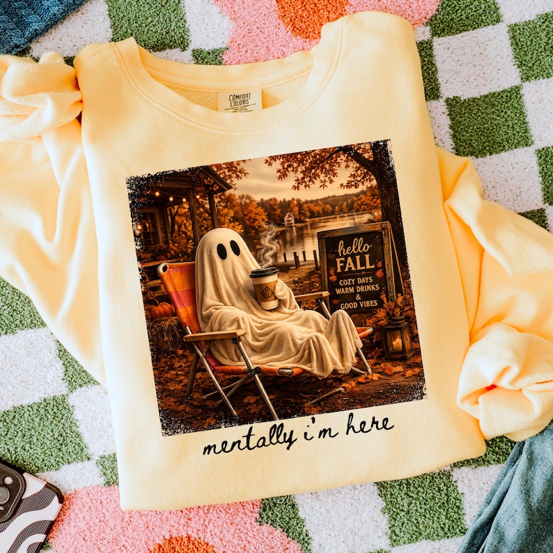 May include: A light yellow sweatshirt with a graphic of a ghost relaxing in a chair, holding a coffee cup, with the text "hello FALL" and "mentally i'm here". The image has a fall theme with a lake and trees.