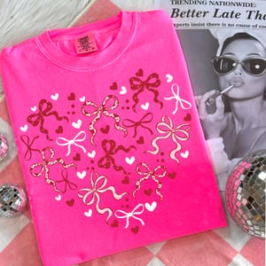 May include: A bright pink sweatshirt with a heart design made of red and white bows and hearts. The shirt has a crew neck and long sleeves. The text on the top right says "TRENDING NATIONWIDE: Better Late The".