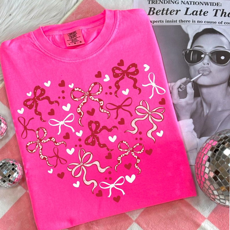 May include: A bright pink sweatshirt with a heart design made of red and white bows and hearts. The shirt has a crew neck and long sleeves. The text on the top right says "TRENDING NATIONWIDE: Better Late The".
