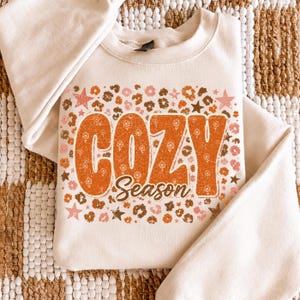 Może przedstawiać: Kremowa bluza z okrągłym dekoltem z dużym nadrukiem graficznym z napisem "COZY Season" w kolorze pomarańczowym. Wzór otoczony jest motywem brązowych i różowych cętek lamparta oraz małymi gwiazdkami, z kwiatową teksturą wewnątrz liter.