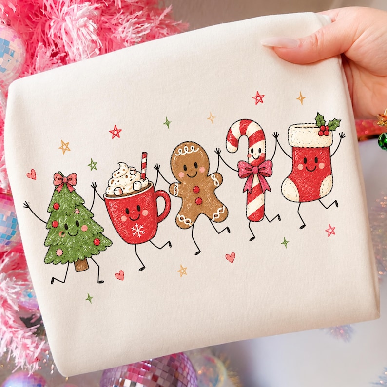 May include: A cream-colored sweatshirt featuring a colorful graphic print of five anthropomorphic holiday characters: a green Christmas tree, a red mug of hot cocoa, a brown gingerbread man, a candy cane, and a red Christmas stocking.