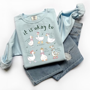 Pode incluir: Um moletom azul claro com a frase "it is okay to" e ilustra&ccedil;&otilde;es de gansos. O moletom &eacute; combinado com jeans azul claro. As ilustra&ccedil;&otilde;es de gansos t&ecirc;m v&aacute;rios acess&oacute;rios e bal&otilde;es de texto.