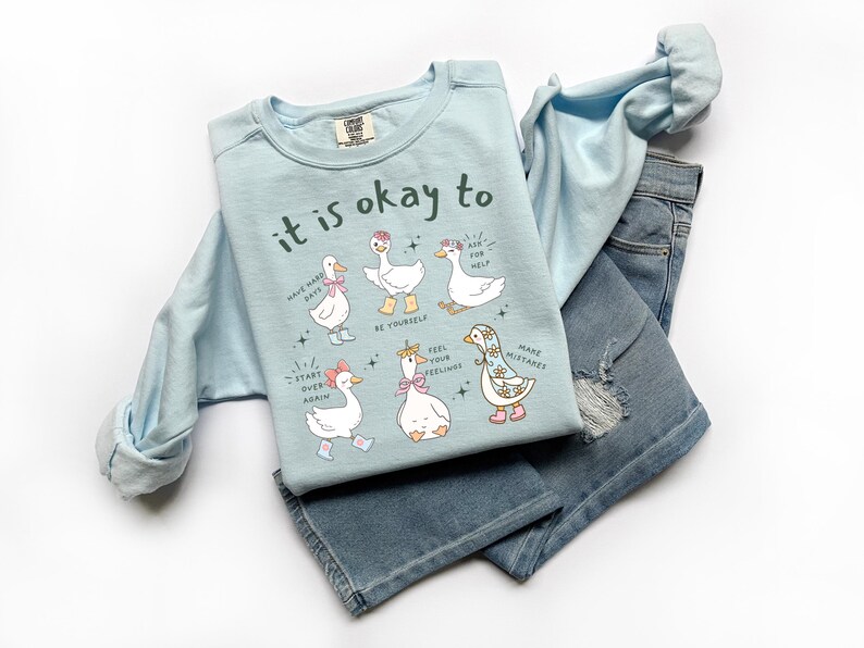 Pode incluir: Um moletom azul claro com a frase "it is okay to" e ilustra&ccedil;&otilde;es de gansos. O moletom &eacute; combinado com jeans azul claro. As ilustra&ccedil;&otilde;es de gansos t&ecirc;m v&aacute;rios acess&oacute;rios e bal&otilde;es de texto.