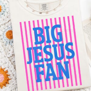 Puede incluir: Camiseta color crema con el texto azul "BIG JESUS FAN" sobre un fondo de rayas rosas verticales. Un delicado collar plateado con peque&ntilde;os abalorios descansa sobre la camiseta. La camiseta es de material Comfort Colors.