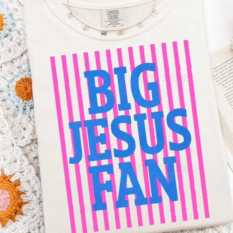 Puede incluir: Camiseta color crema con el texto azul "BIG JESUS FAN" sobre un fondo de rayas rosas verticales. Un delicado collar plateado con peque&ntilde;os abalorios descansa sobre la camiseta. La camiseta es de material Comfort Colors.