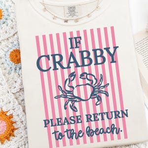 Puede incluir: Camiseta color crema con el texto "IF CRABBY PLEASE RETURN to the beach." El texto es azul marino y el fondo tiene rayas verticales rosas. Un gr&aacute;fico de cangrejo azul marino est&aacute; centrado debajo de la palabra "CRABBY". Un delicado collar de mariposas est&aacute; en la parte superior.