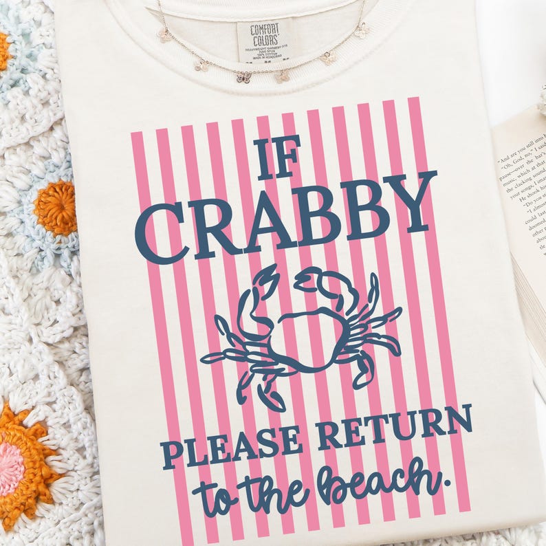 Puede incluir: Camiseta color crema con el texto "IF CRABBY PLEASE RETURN to the beach." El texto es azul marino y el fondo tiene rayas verticales rosas. Un gr&aacute;fico de cangrejo azul marino est&aacute; centrado debajo de la palabra "CRABBY". Un delicado collar de mariposas est&aacute; en la parte superior.