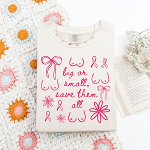 May include: Cream-colored t-shirt with pink graphic text and illustrations. The text reads "big or small, save them all" with drawings of bows, breasts, and ribbons. A crochet blanket and open book are in the background.