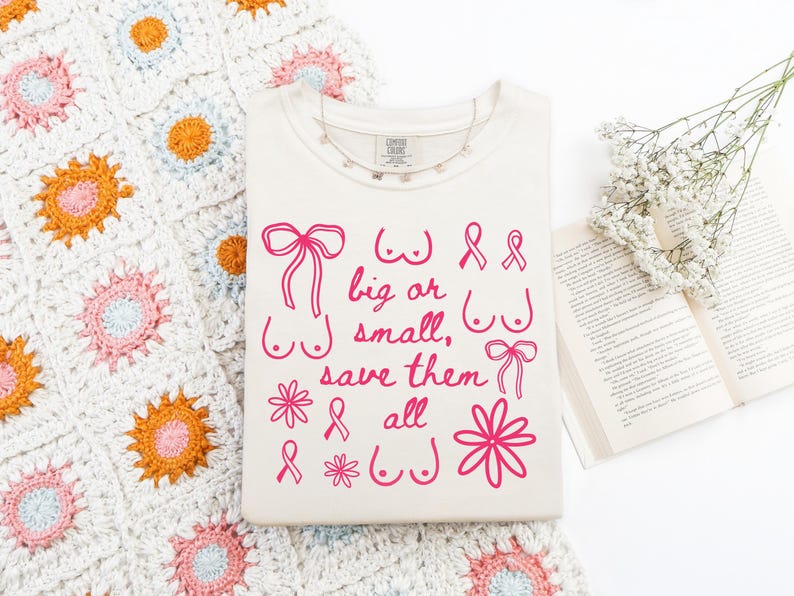 May include: Cream-colored t-shirt with pink graphic text and illustrations. The text reads "big or small, save them all" with drawings of bows, breasts, and ribbons. A crochet blanket and open book are in the background.
