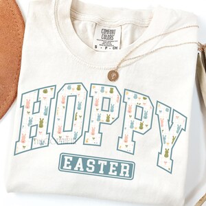 May include: Cream-colored t-shirt with the words "HOPPY EASTER" in a blue and white design. The word "HOPPY" is filled with a pattern of pastel-colored bunnies. A gold necklace with a pendant rests on the shirt.