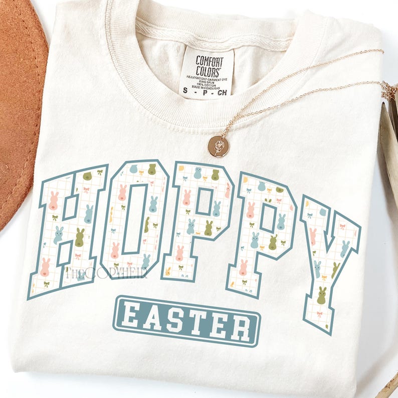 May include: Cream-colored t-shirt with the words "HOPPY EASTER" in a blue and white design. The word "HOPPY" is filled with a pattern of pastel-colored bunnies. A gold necklace with a pendant rests on the shirt.