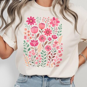 May include: Cream-colored t-shirt featuring a vibrant floral design. The graphic showcases a variety of pink flowers, including tulips and daisies, with green leaves and stems. The design is centered on the shirt, creating a visually appealing botanical theme.