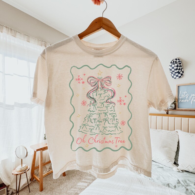 Peut inclure: T-shirt cr&egrave;me avec un motif de sapin de No&euml;l. L'arbre est compos&eacute; de n&oelig;uds et de rubans roses et verts, encadr&eacute; par une bordure verte avec des flocons de neige et des &eacute;toiles. Le texte "Oh Christmas Tree" est en dessous.