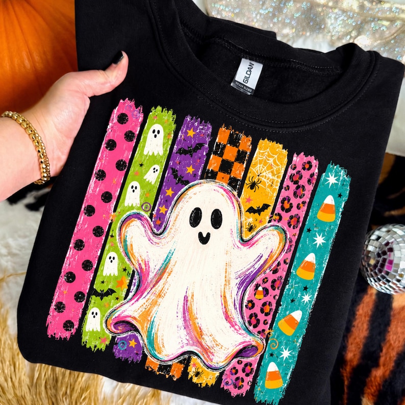 May include: A black crewneck sweatshirt featuring a central white ghost graphic layered over vertical brushstroke stripes. The stripes display patterns including polka dots, ghosts, bats, orange checkers, spiderwebs, leopard print, and candy corn.