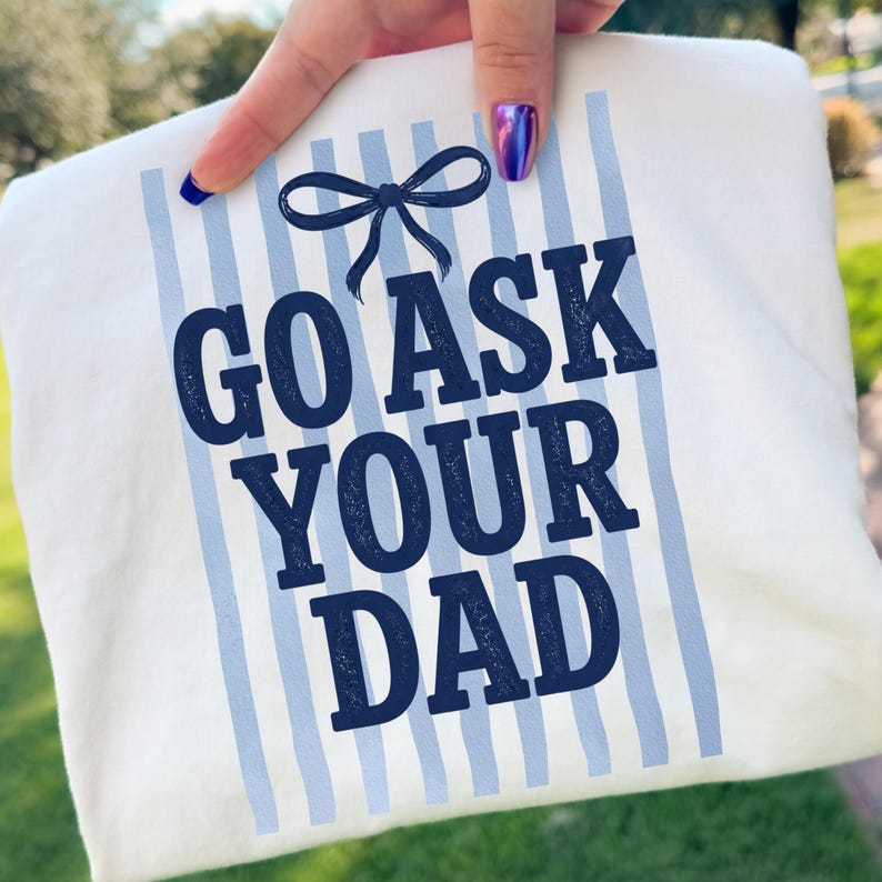 Puede incluir: Camiseta blanca con rayas verticales azules y el texto "GO ASK YOUR DAD" en azul marino. Un lazo azul marino est&aacute; encima del texto. La camiseta est&aacute; sujeta por una mano con esmalte de u&ntilde;as azul.