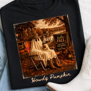 Può includere: T-shirt nero con grafica a tema autunnale. La grafica raffigura un fantasma con cappello e stivali da cowboy, seduto su una sedia da campeggio, con in mano una tazza di caffè, con la scritta "hello FALL" e "Howdy Pumpkin".