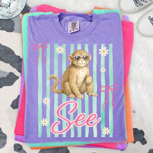 May include: Stacked t-shirts in shades of purple, pink, orange, and green. The top shirt is purple with a graphic of a monkey wearing sunglasses, daisies, and the word "See". The shirt has vertical stripes and bows.