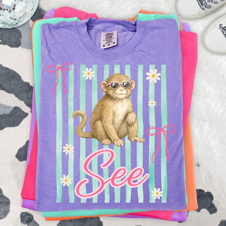 May include: Stacked t-shirts in shades of purple, pink, orange, and green. The top shirt is purple with a graphic of a monkey wearing sunglasses, daisies, and the word "See". The shirt has vertical stripes and bows.