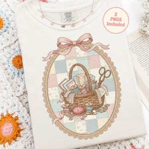 May include: Cream-colored sweatshirt with a detailed illustration of a sewing basket filled with spools of thread, fabric scraps, and scissors. The design is framed by a pink ribbon and a decorative oval border. The text "2 PNGs Included" is in a pink circle.