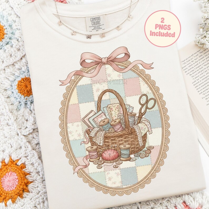May include: Cream-colored sweatshirt with a detailed illustration of a sewing basket filled with spools of thread, fabric scraps, and scissors. The design is framed by a pink ribbon and a decorative oval border. The text "2 PNGs Included" is in a pink circle.