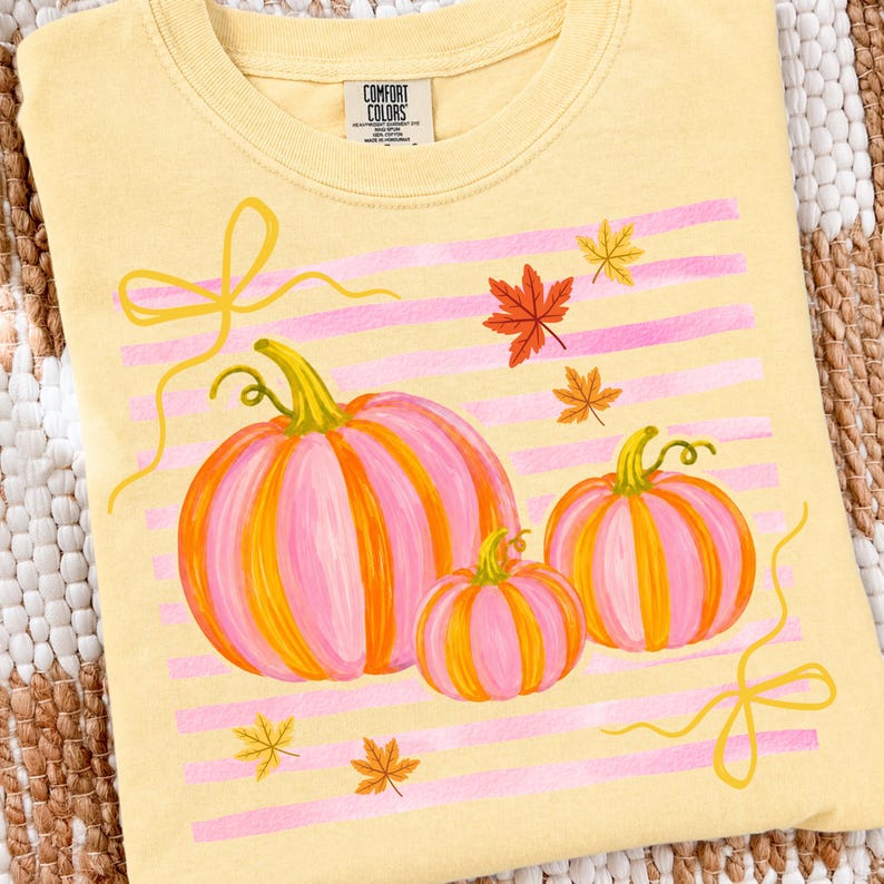 May include: A light yellow t-shirt featuring a design of three pink and orange pumpkins with green stems. Pink horizontal stripes and fall leaves are also included in the design. The shirt has the text "COMFORT COLORS" on the tag.