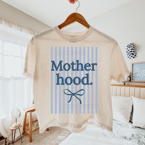 K&ouml;nnte beinhalten: Cremefarbenes T-Shirt mit dem Wort "Motherhood" in Marineblau, vor einem hellblau gestreiften Hintergrund. Eine marineblaue Schleifengrafik befindet sich unter dem Text. Der Markenname "THECOPTIER" ist oben und unten auf dem Design aufgedruckt.