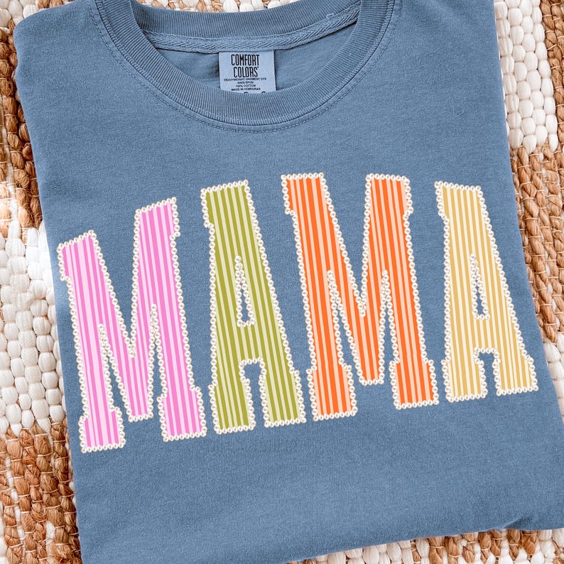 Puede incluir: Una camiseta azul Comfort Colors con la palabra "MAMA" en letras a rayas de colores. Las letras son rosas, verdes, naranjas y amarillas con un contorno blanco. La camiseta tiene cuello redondo.