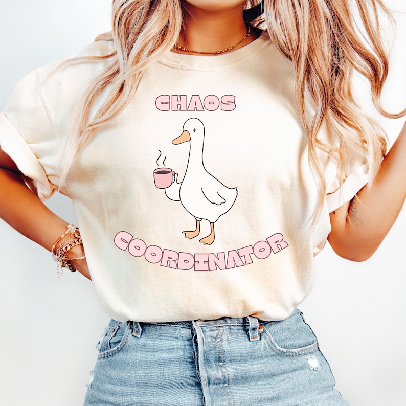 May include: Cream-colored t-shirt featuring a cartoon duck holding a pink mug. The text "CHAOS COORDINATOR" is printed above and below the duck in a pink, rounded font. The duck is white with a yellow beak and feet.