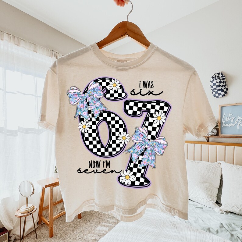 Peut inclure: T-shirt beige avec un motif graphique repr&eacute;sentant le chiffre 67 en damier noir et blanc, orn&eacute; de n&oelig;uds floraux et de marguerites. Le texte "I WAS SIX NOW I'M seven" est &eacute;galement imprim&eacute;. Le t-shirt est suspendu &agrave; un cintre en bois.